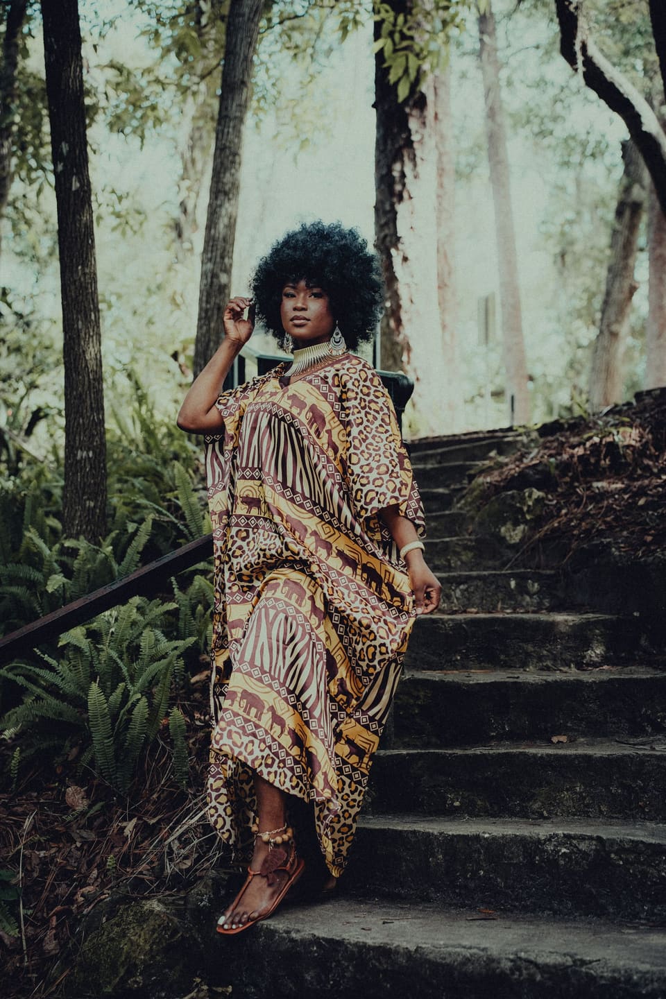 Model in a vibrant patterned kaftan posing on stone steps in a lush forest environment.