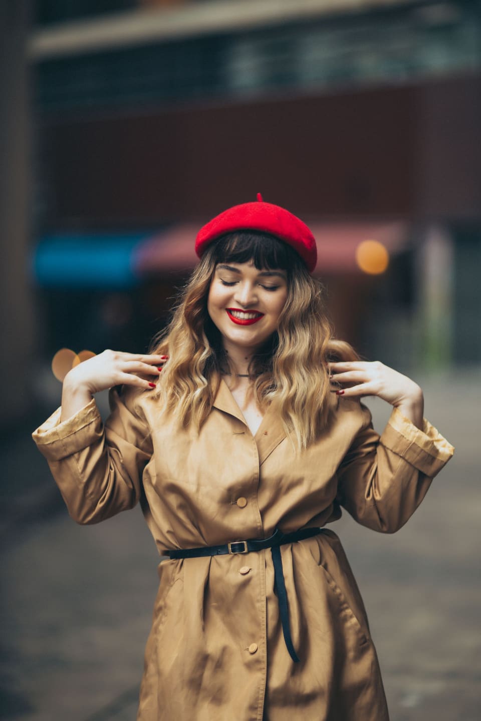 Parisian chic: Fashionable woman in red beret and trench coat posing outdoors with a smile.
