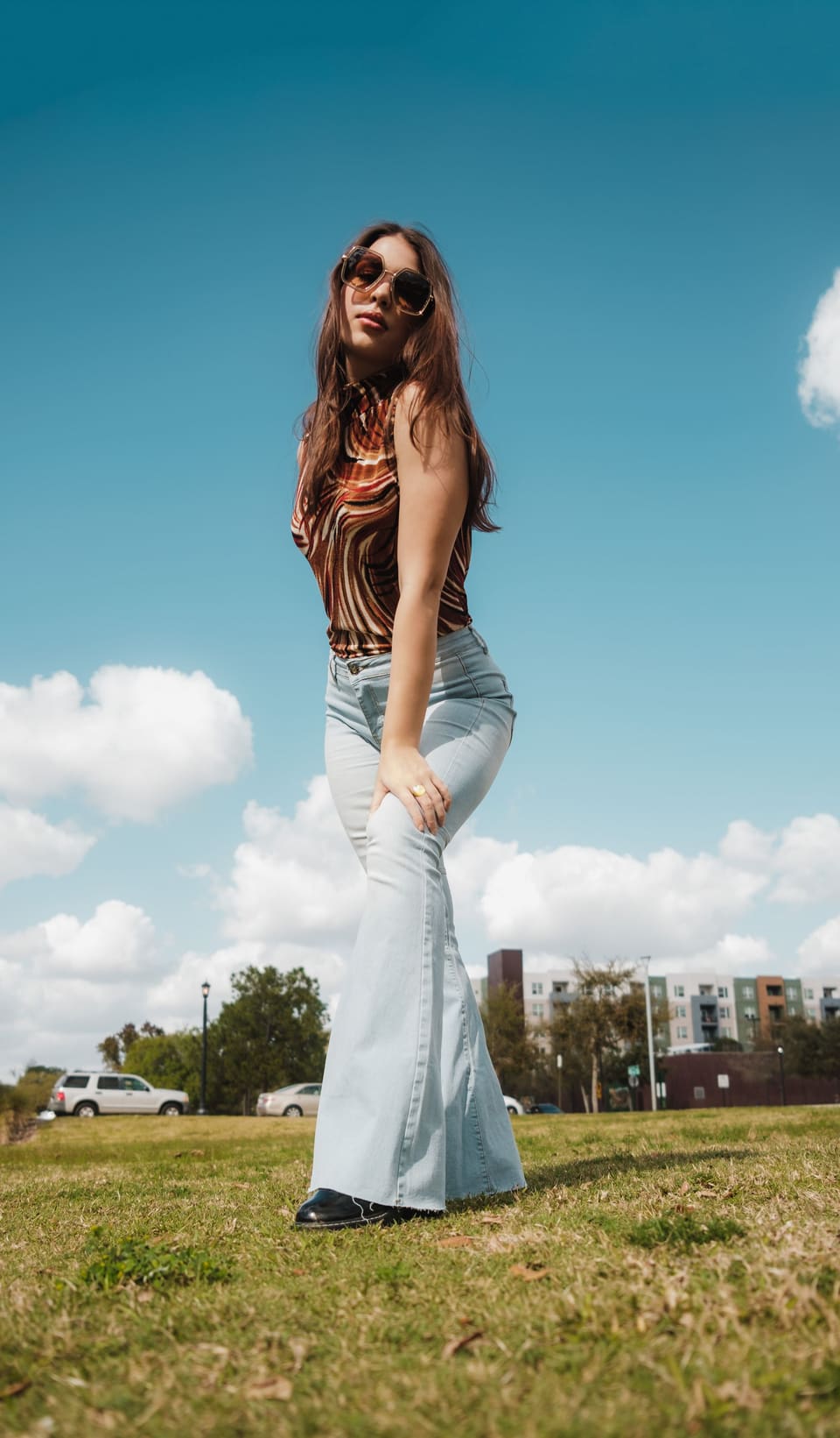 Fashionable woman with sunglasses poses in a park on a sunny day, showcasing retro flared jeans.