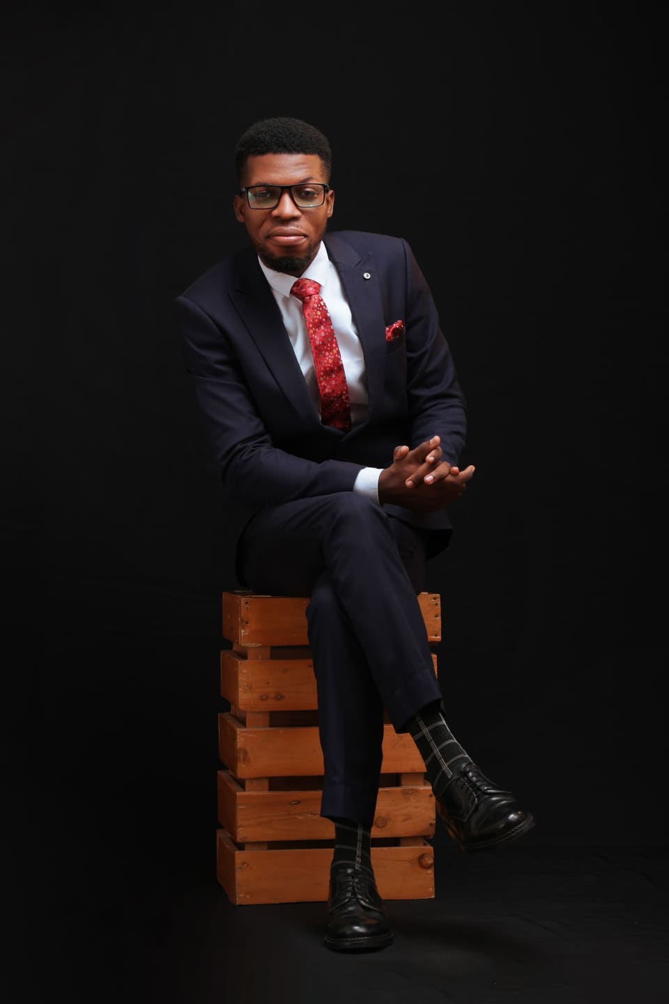 Stylish man in formal navy suit and red tie posing confidently on a wooden crate.