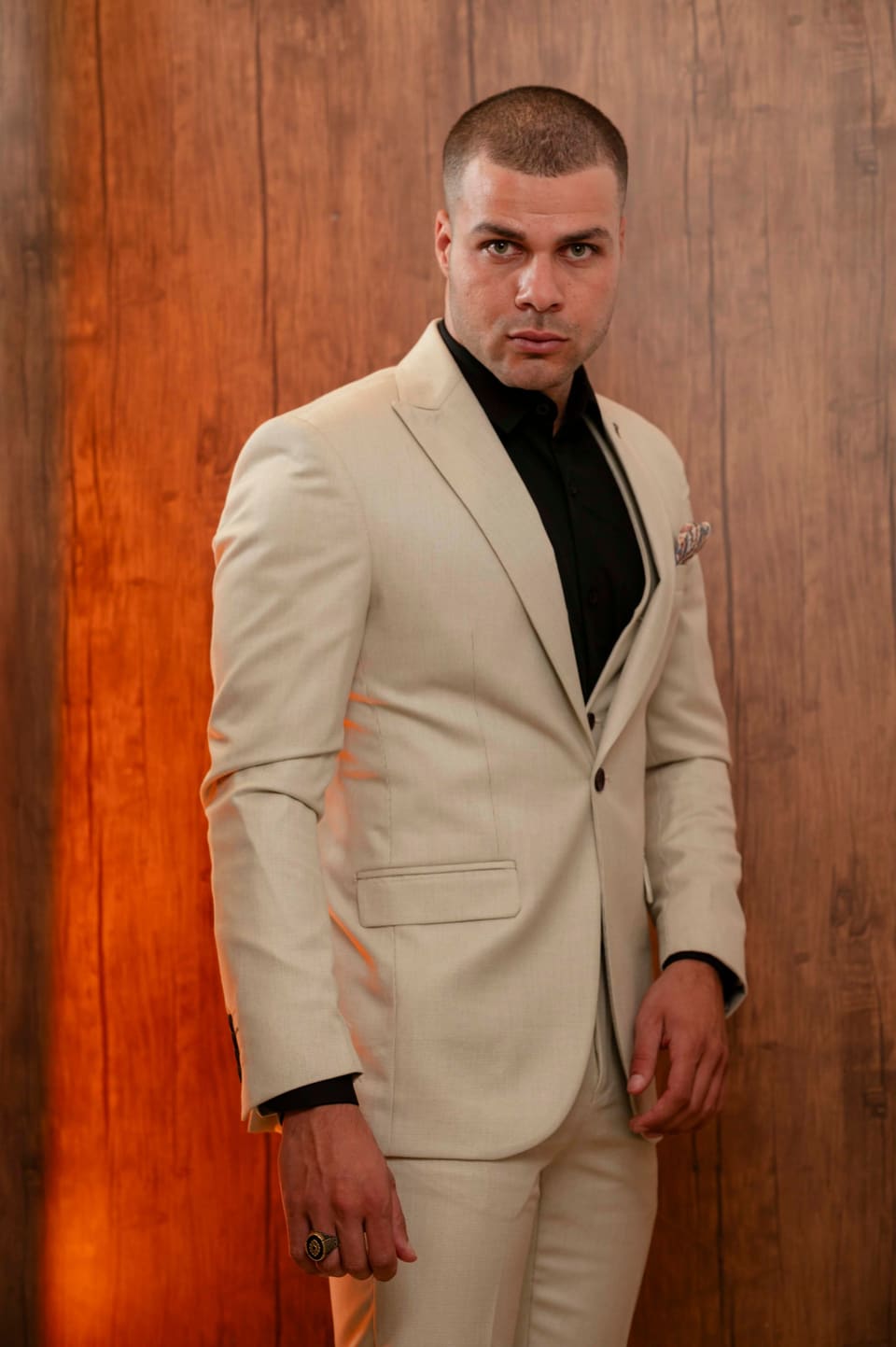 Confident man posing indoors in a beige suit and black shirt against a warm backdrop.