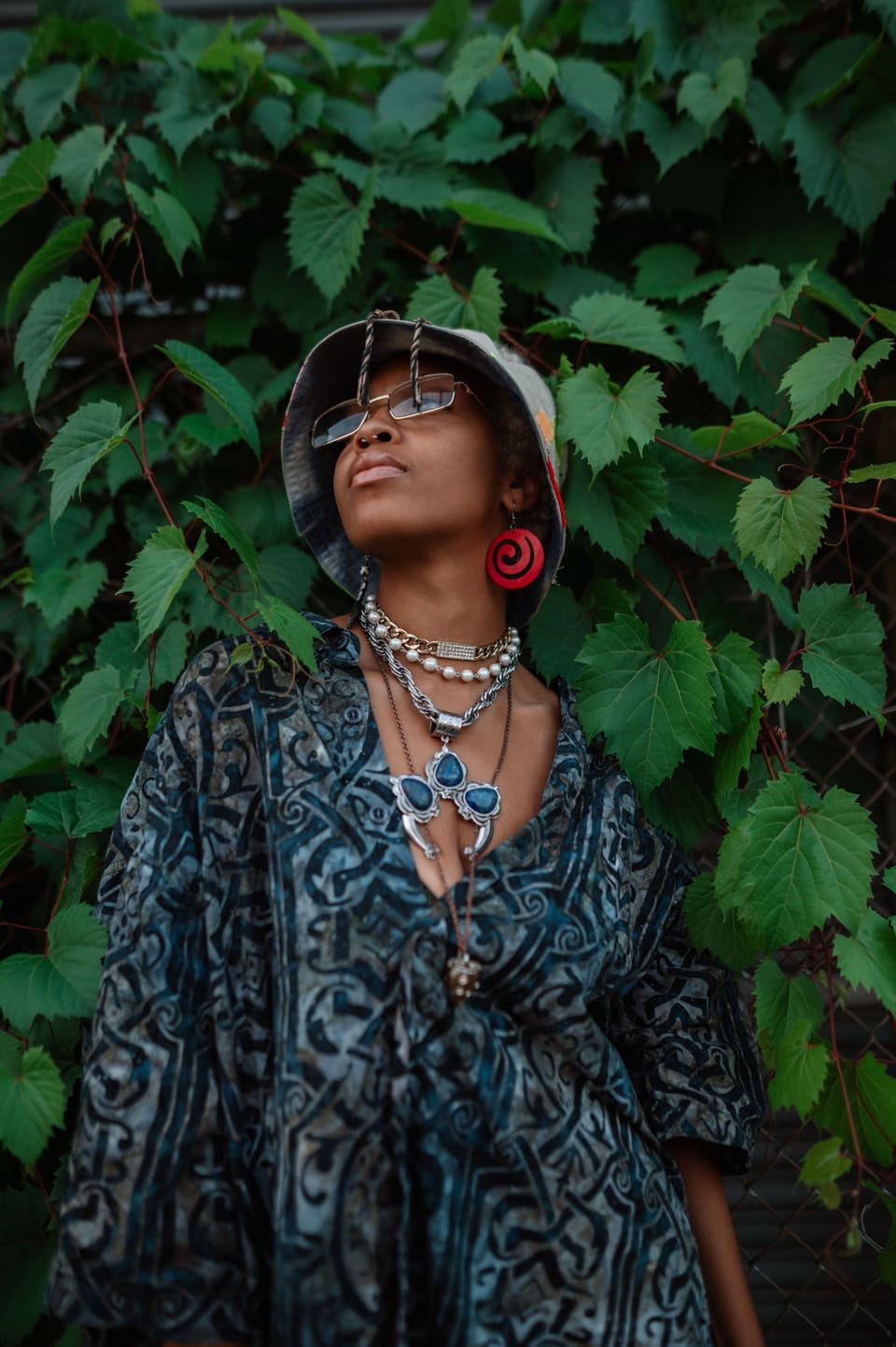 Fashionable woman wearing eyeglasses and hat poses against lush green backdrop.