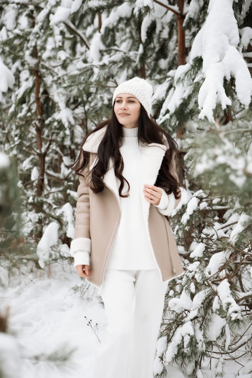 Cosy knits: A young woman in stylish winter clothing poses happily amidst snowy conifer trees.