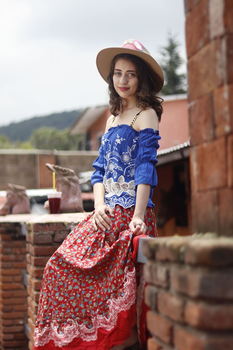 A stylish young woman poses in bohemian clothing against a rustic outdoor backdrop.