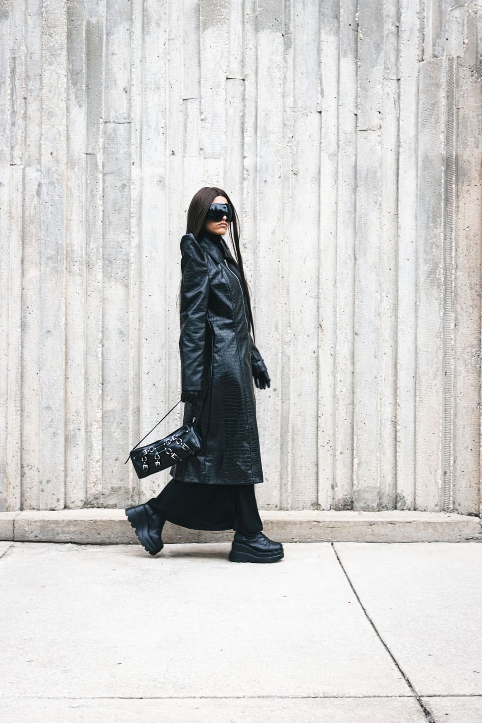 Downtown all-black: Stylish woman walking with black leather trench coat and sunglasses against concrete wall.