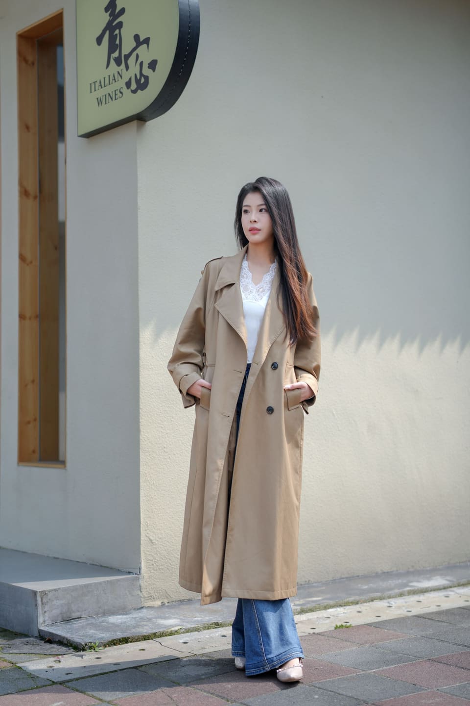 Seoul layering: Stylish young woman posing in a trench coat outside an Italian wine shop.