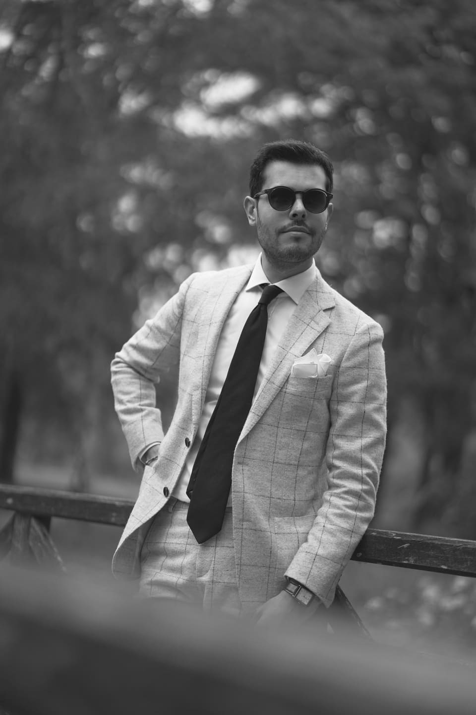 A fashionable man in a suit and sunglasses poses confidently in a park setting.