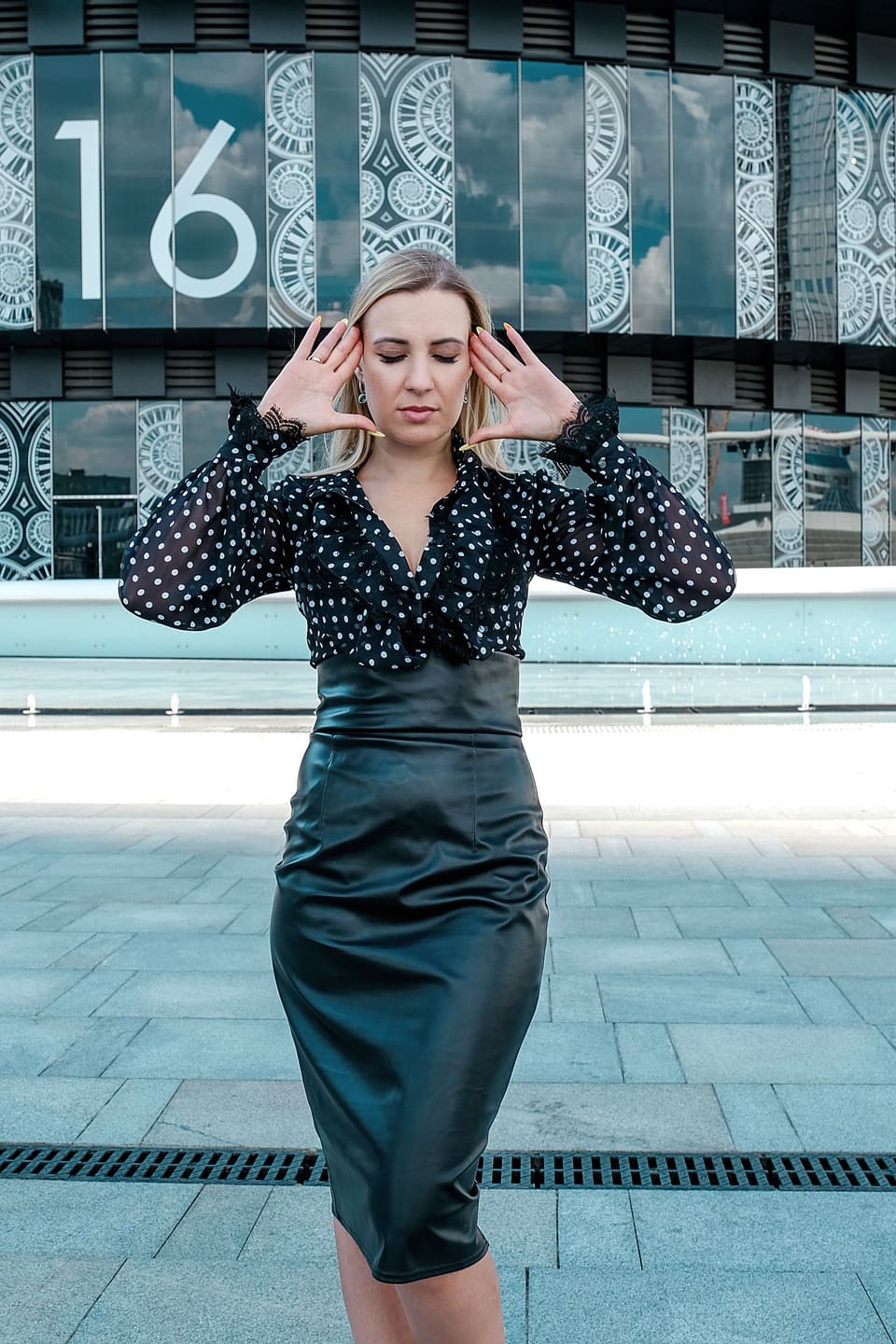 Office siren: Elegant young woman in black leather skirt touching head while standing with eyes closed outside contemporary building in daylight